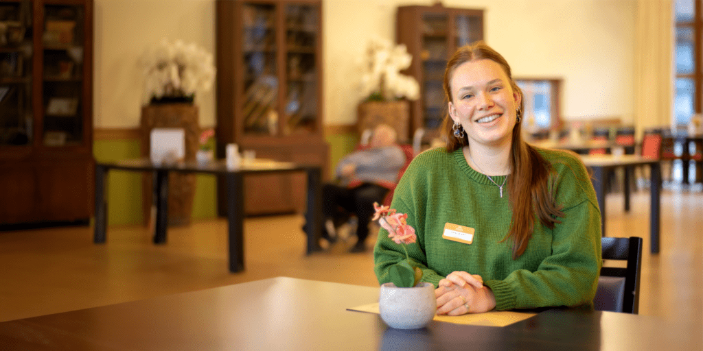 Een jonge vrouw met lang roodbruin haar en een groene trui zit glimlachend aan een tafel in een gezellige binnenruimte. Voor haar staat een kleine bloempot met een roze bloem. Op de achtergrond zijn enkele meubels en een oudere persoon in een rolstoel zichtbaar, die een warme sfeer suggereren.